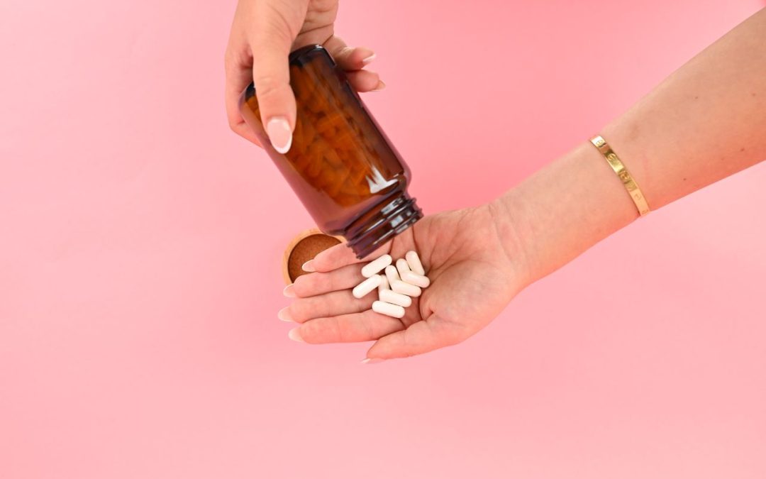 Une personne versant des gélules blanches d'un flacon en verre ambré dans sa main sur fond rose.